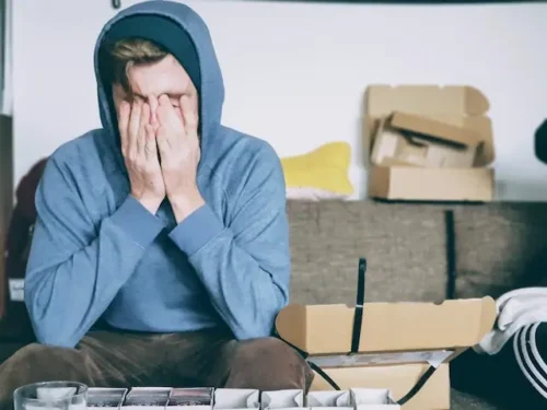 Man wearing a blue hoodie sitting on a couch covered in cardboard boxes