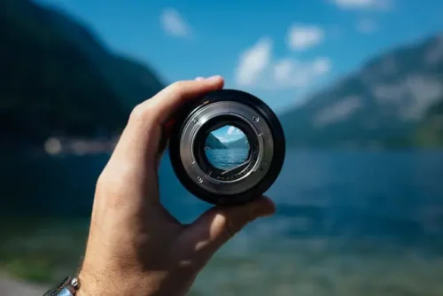 A person holding a camera lens with a nature background.