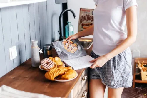 A woman reading a cookbook on a table with a plate of pastries.