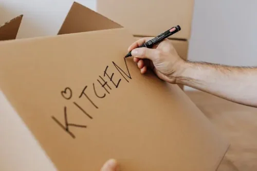 A person labeling a cardboard box 'kitchen.'