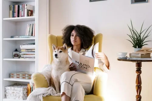 Woman in pajamas reading a magazine with her dog beside her in a cozy yellow armchair.