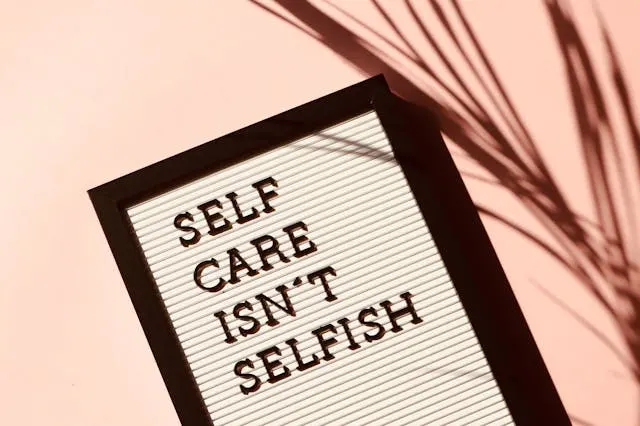 Letter board reading “SELF CARE ISN’T SELFISH” on a pink surface with palm shadow, representing wellness rituals to reset